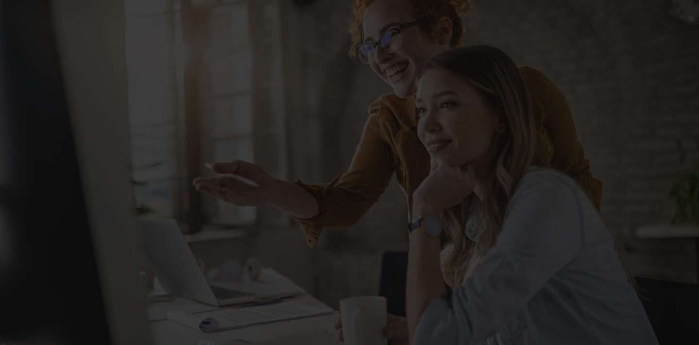 Two Women Smilingly Stare at a Computer Screen.