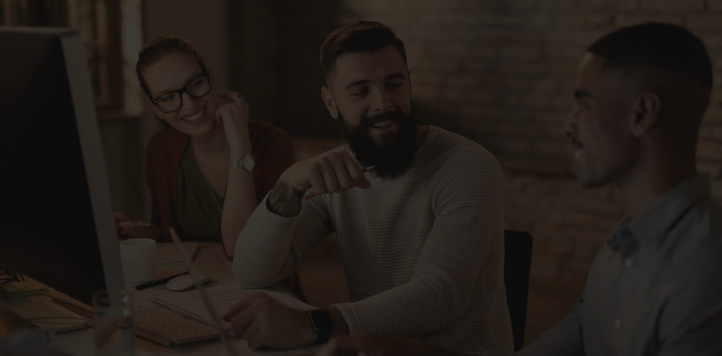 Three Individuals Smiling and Talking While Sitting at a Desk With Computers.