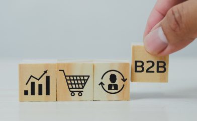 Three Wooden Blocks Arranged in a Row on a Light Surface Showing Icons and A Hand Placing a Fourth Wooden Block Labeled 'B2B'.