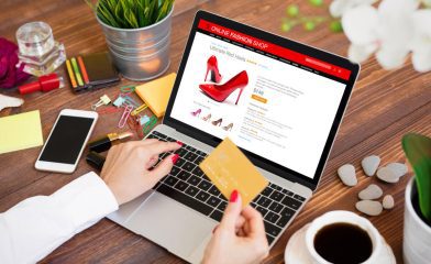 An Open Laptop on a Wooden Desk Displaying an Online Fashion Shop Page With a Prominent Image of Red High-Heeled Shoes. A Person Holding a Gold Credit Card While their Other Hand Navigates the Laptop's Trackpad.