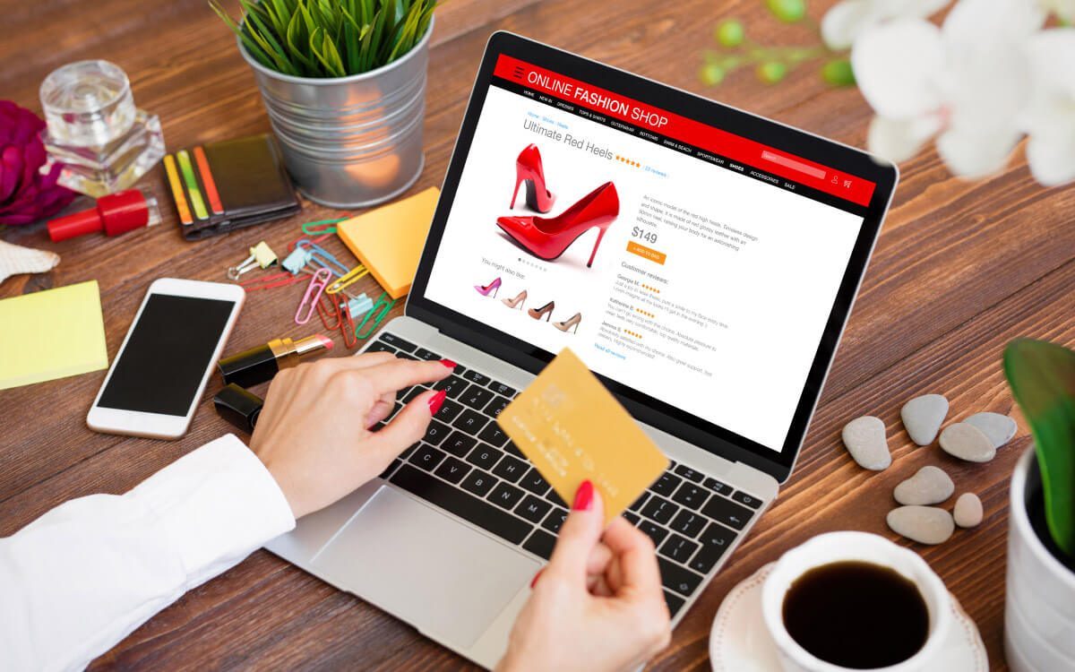 An Open Laptop on a Wooden Desk Displaying an Online Fashion Shop Page With a Prominent Image of Red High-Heeled Shoes. A Person Holding a Gold Credit Card While their Other Hand Navigates the Laptop's Trackpad.