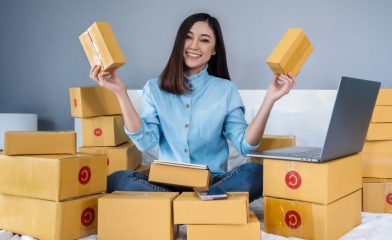 A Smiling Woman Sitting on a Bed Surrounded by Numerous Cardboard Boxes, A Smartphone, and a Laptop.