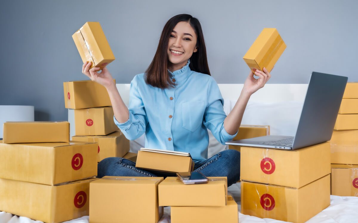 A Smiling Woman Sitting on a Bed Surrounded by Numerous Cardboard Boxes, A Smartphone, and a Laptop.