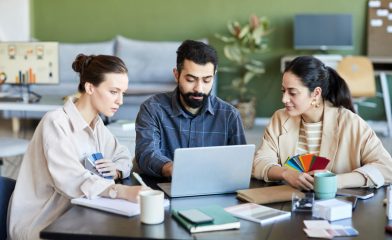 Three Individuals Seated at a Table in a Vibrant Office Environment, Facing a Laptop.