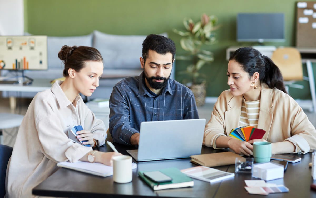 Three Individuals Seated at a Table in a Vibrant Office Environment, Facing a Laptop.