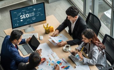 Three Individuals in a Professional Setting Engaged in Work-Related Activities On A Desk With a Large Computer Monitor Displaying Graphics Related to SEO (Search Engine Optimization).