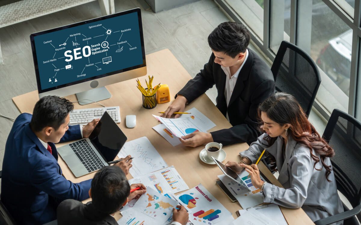Three Individuals in a Professional Setting Engaged in Work-Related Activities On A Desk With a Large Computer Monitor Displaying Graphics Related to SEO (Search Engine Optimization).
