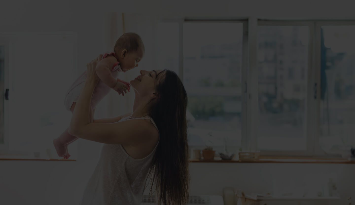 A Smiling Woman Holding a Baby Up in the Air Indoors, With a Window Showing a Cityscape in the Background.