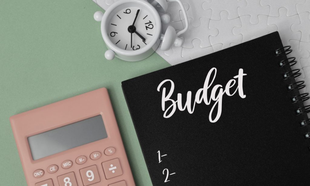 Black notebook with "Budget" written on it, pink calculator, white clock, and puzzle pieces on green and white background.