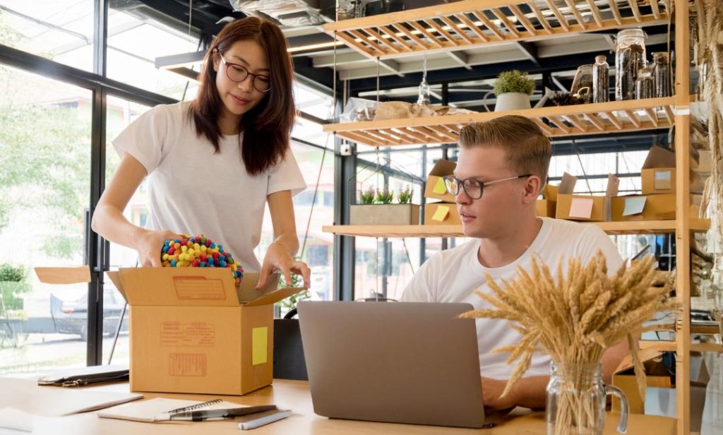 A woman packs colorful items into a cardboard box while a man works on a laptop beside her, symbolizing eCommerce entrepreneurship.