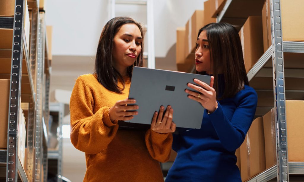 Two women in a warehouse discuss work while looking at a laptop.