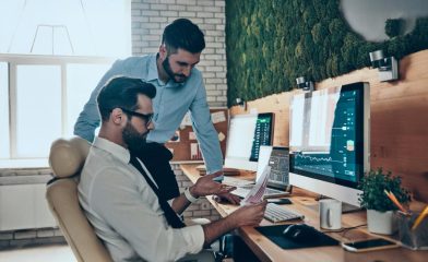 Two professionals in an office reviewing documents with data charts on monitors.