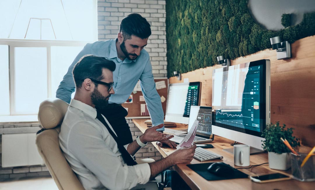 Two professionals in an office reviewing documents with data charts on monitors.