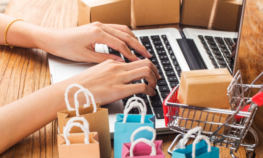 A person types on a laptop surrounded by mini shopping bags, boxes, and a small shopping cart, symbolizing eCommerce.
