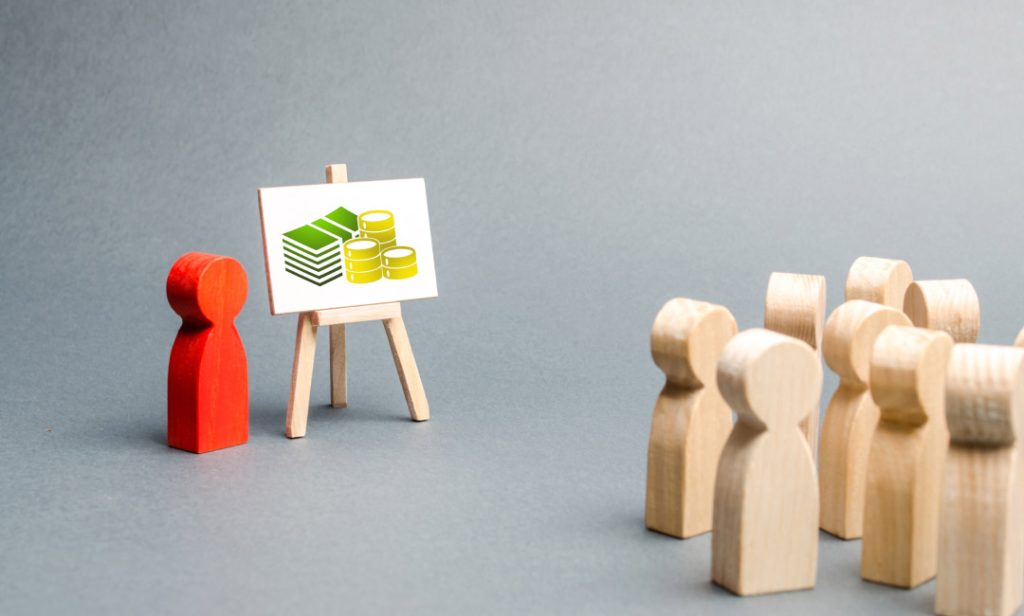 Wooden figurines in a presentation on funding and investment, with cash and coins displayed on a board.