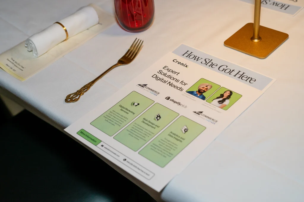 Close-up of a Cronix flyer at a table at the &ldquo;How She Got Here&rdquo; event, featuring a red glass and neatly rolled napkin on a white tablecloth.
