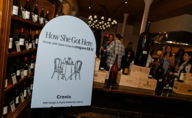 An event sign reading &ldquo;How She Got Here &ndash; Dinner with Cass Cross & empowHER&rdquo; stands in front of wine bottles and mingling guests at Italian Wine Merchants, NYC.