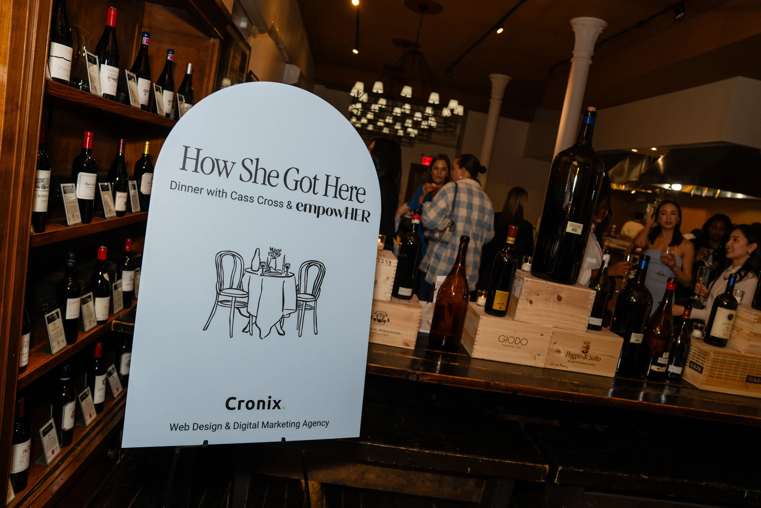 An event sign reading &ldquo;How She Got Here &ndash; Dinner with Cass Cross & empowHER&rdquo; stands in front of wine bottles and mingling guests at Italian Wine Merchants, NYC.