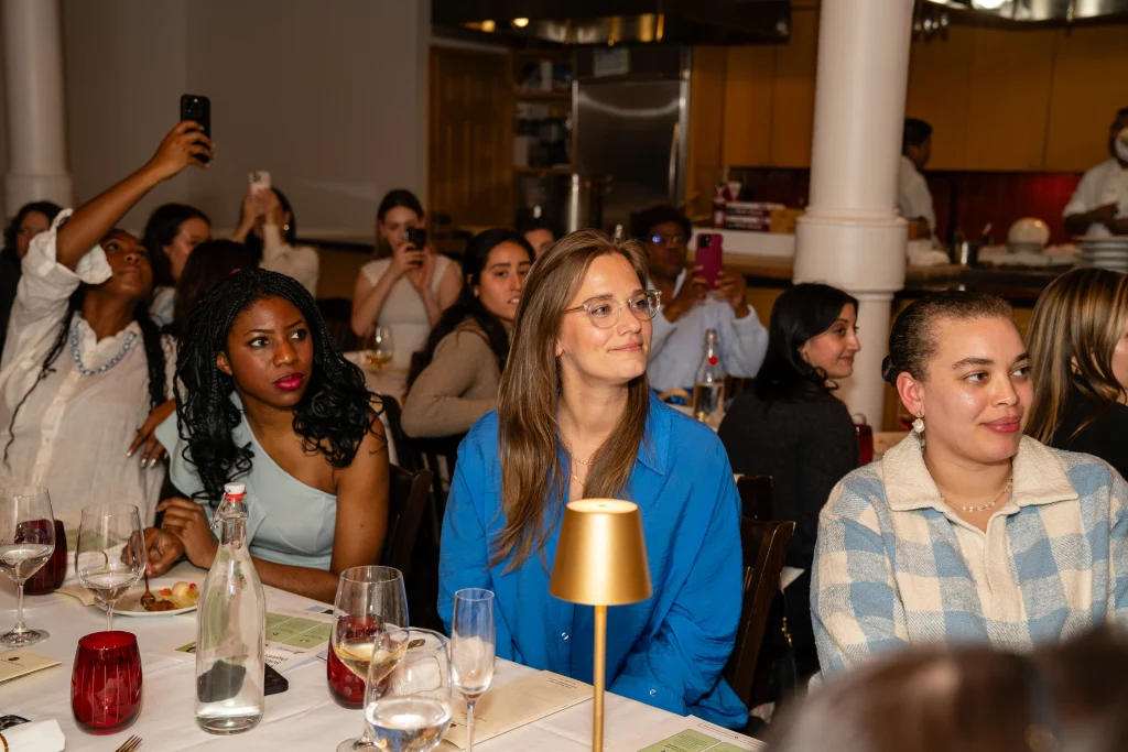 HSGH Season 1 guests Joanna Smith, Sandra Bellows, and Alexandra Felton sit attentively at the &ldquo;How She Got Here&rdquo; dinner event, surrounded by wine glasses and warm ambient lighting as others capture the moment behind them.