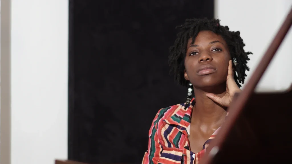 Joanna Smith, wearing a colorful striped shirt, sits thoughtfully with her hand on her face, framed by a dark background and a piano in the foreground.
